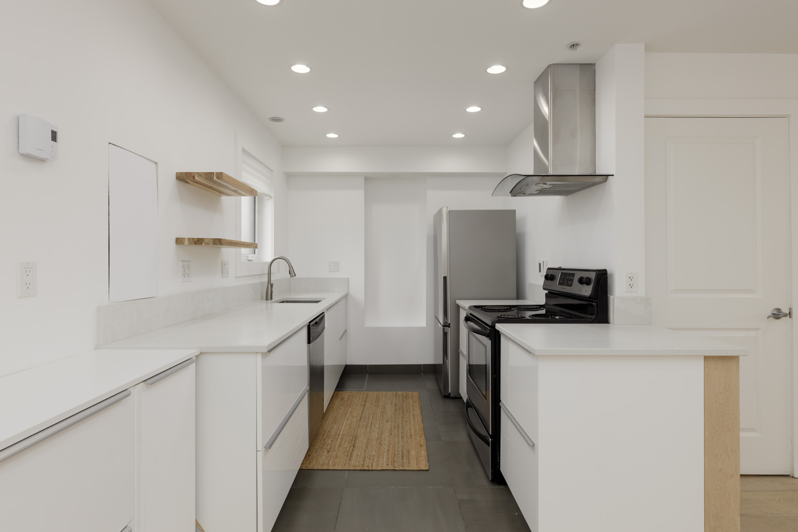 White kitchen with stainless appliances, open shelves, recessed lights, and a beige rug on gray floor in a Vancouver home.