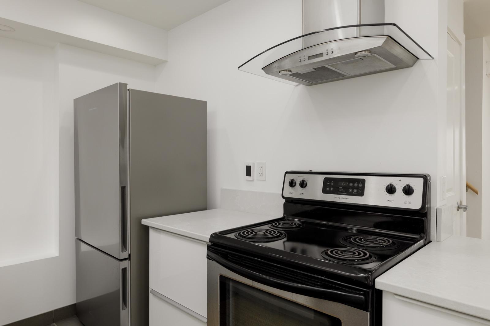 Modern kitchen with stainless steel fridge, electric stove, range hood, white cabinets, and light counters in Vancouver.