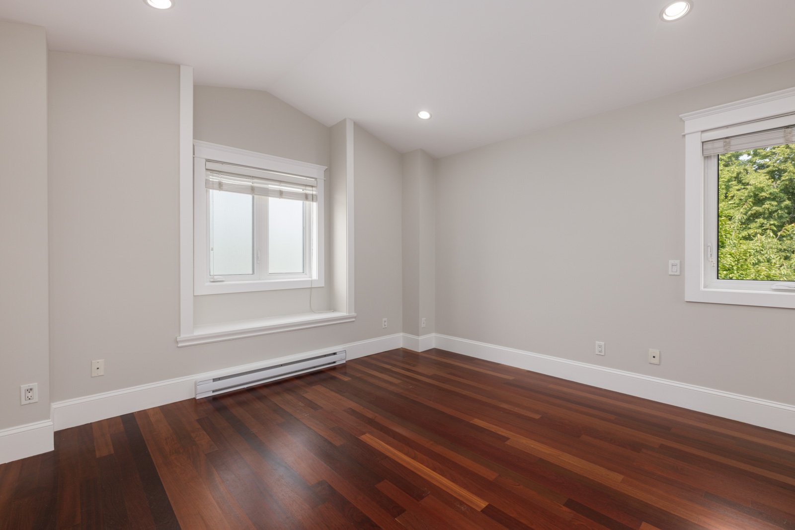 Empty room with light gray walls, two windows, white trim, recessed ceiling lights, and dark wood floors in Vancouver.