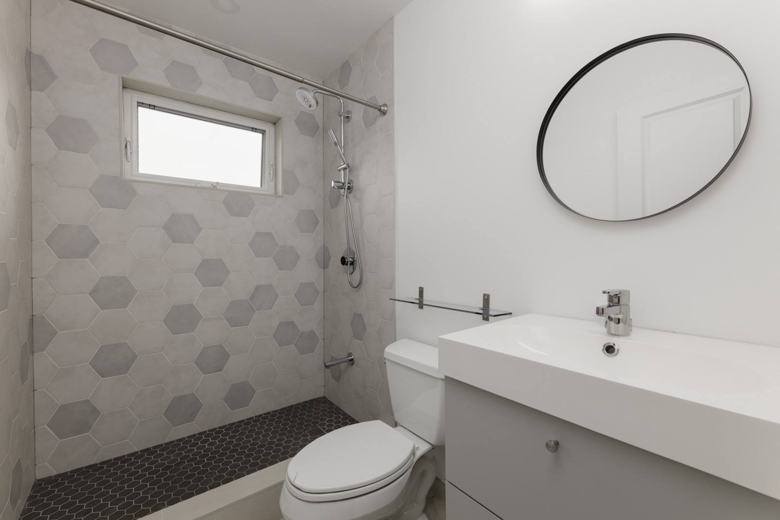 Modern bathroom in Vancouver near St Catherines St and E 8th Ave with hex tile shower, round mirror, white sink, toilet, and shower window.