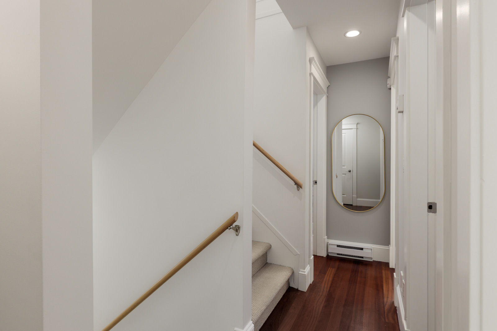 Vancouver hallway with wood floors, white walls, wooden-railed staircase, and a large oval mirror on a gray accent wall at the end.