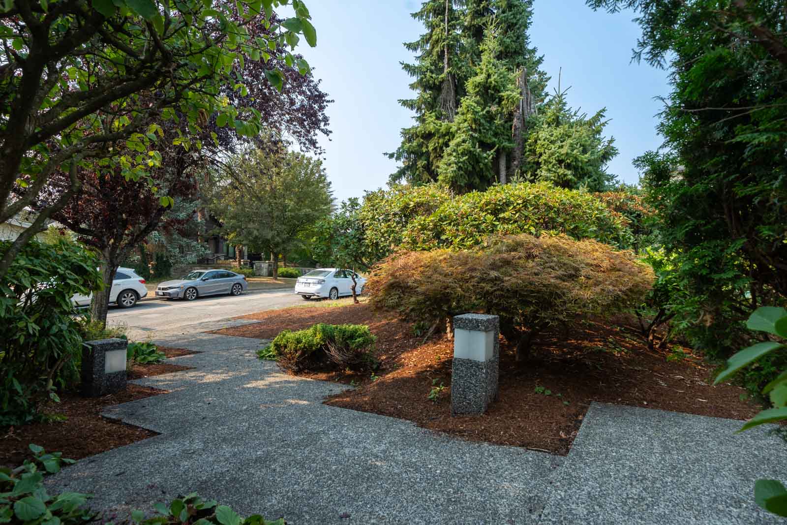 Suburban sidewalk and street with parked cars, trees, bushes, and two stone lamp posts on Vancouver’s W 50th Ave near Ash St.