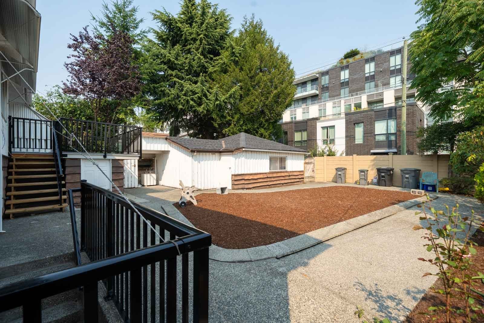 Spacious Vancouver backyard with mulch, concrete patio, small white building, trees, and multi-story apartment in background.