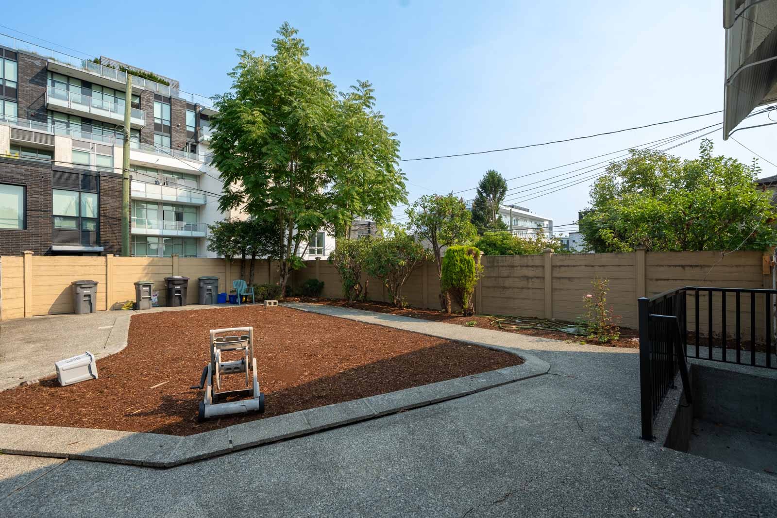 Fenced backyard near W 50th Ave, Vancouver, with mulch, lawn roller, concrete paths, and trash bins; homes in background.