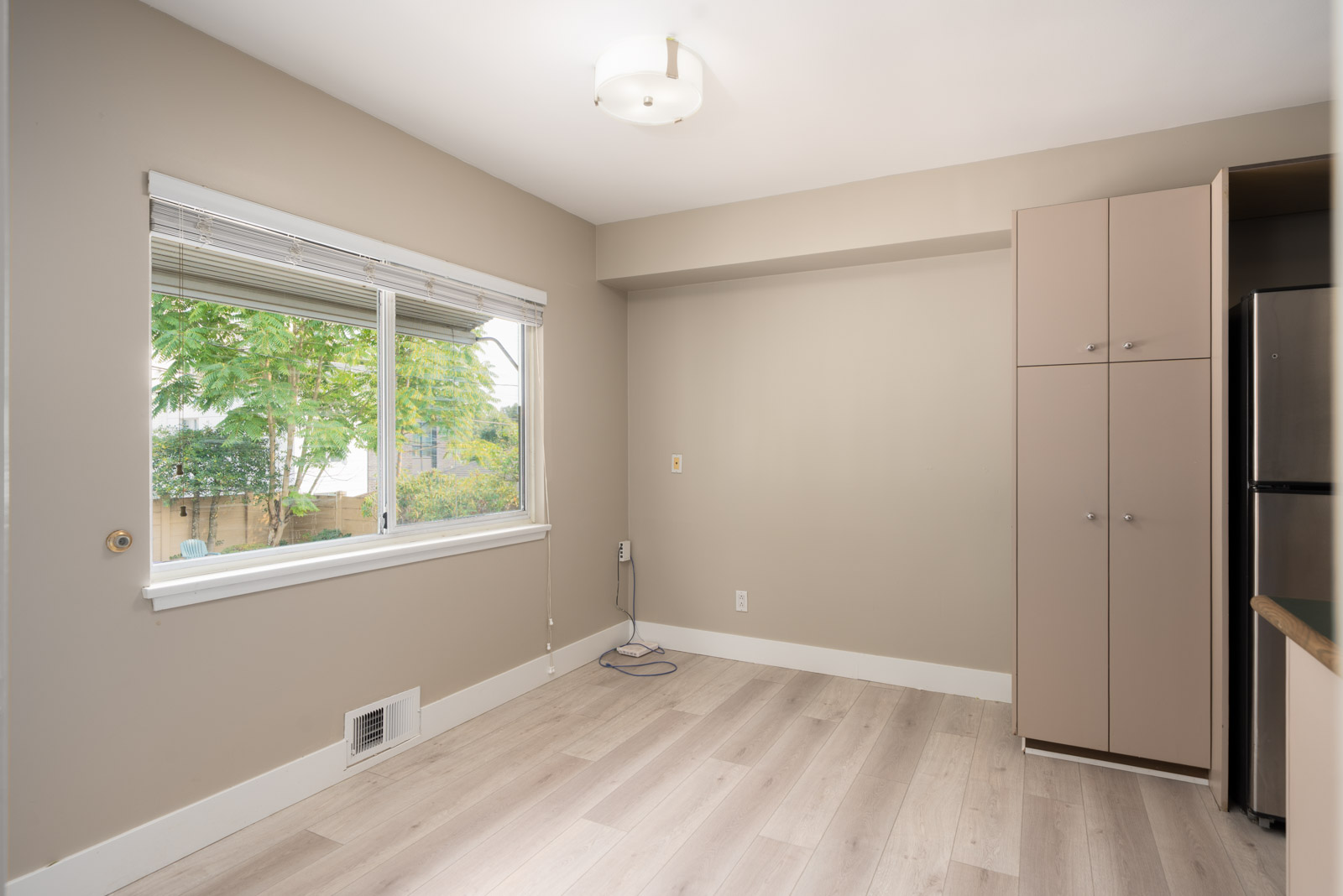 Empty room with light wood floors, beige walls, window blinds, ceiling light, cabinet and fridge; near Ash St & W 50th, Vancouver.