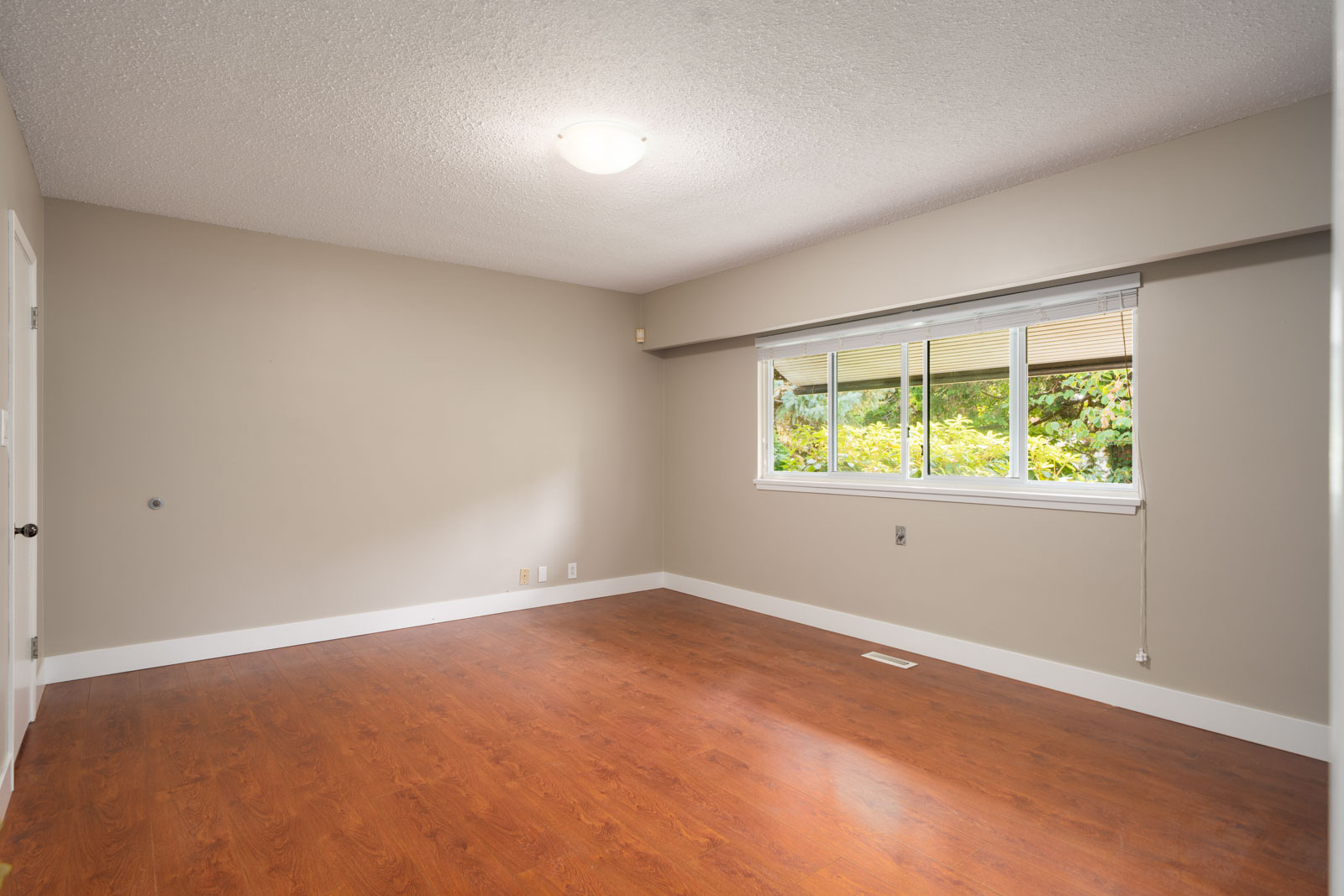 Empty room in Vancouver with beige walls, white trim, wood flooring, large window with shade, and a ceiling light fixture.