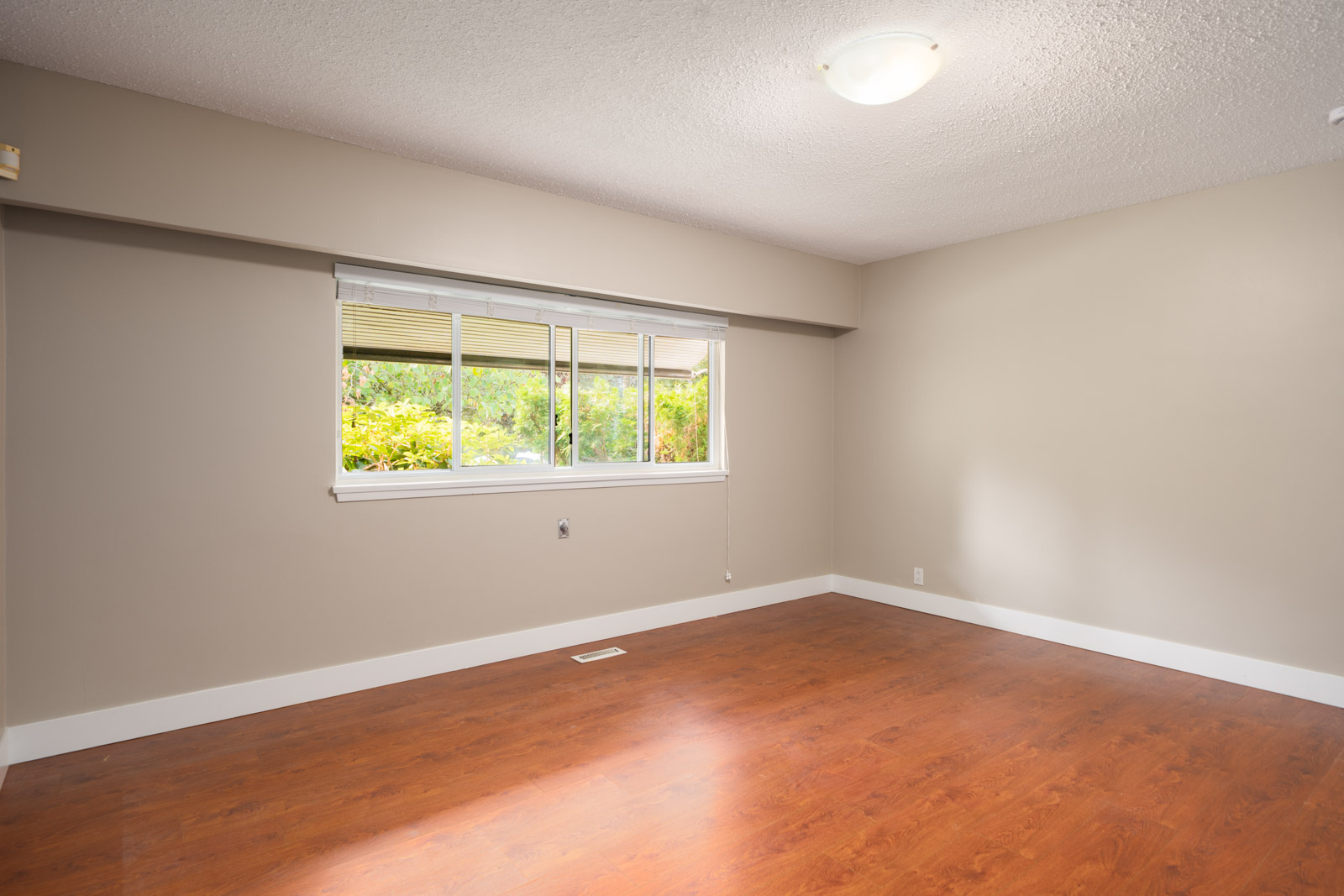 Empty room in Vancouver with beige walls, white trim, wood floor, ceiling light, and a large window overlooking greenery.