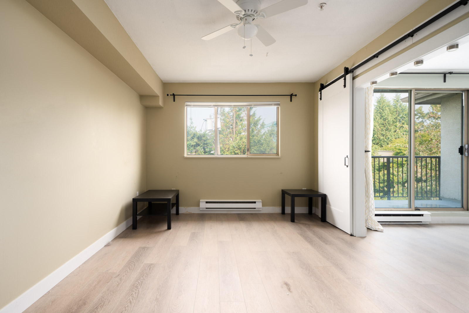 Empty room with light wood floors, beige walls, two black tables, ceiling fan, window, and sliding door to balcony with greenery in Vancouver.
