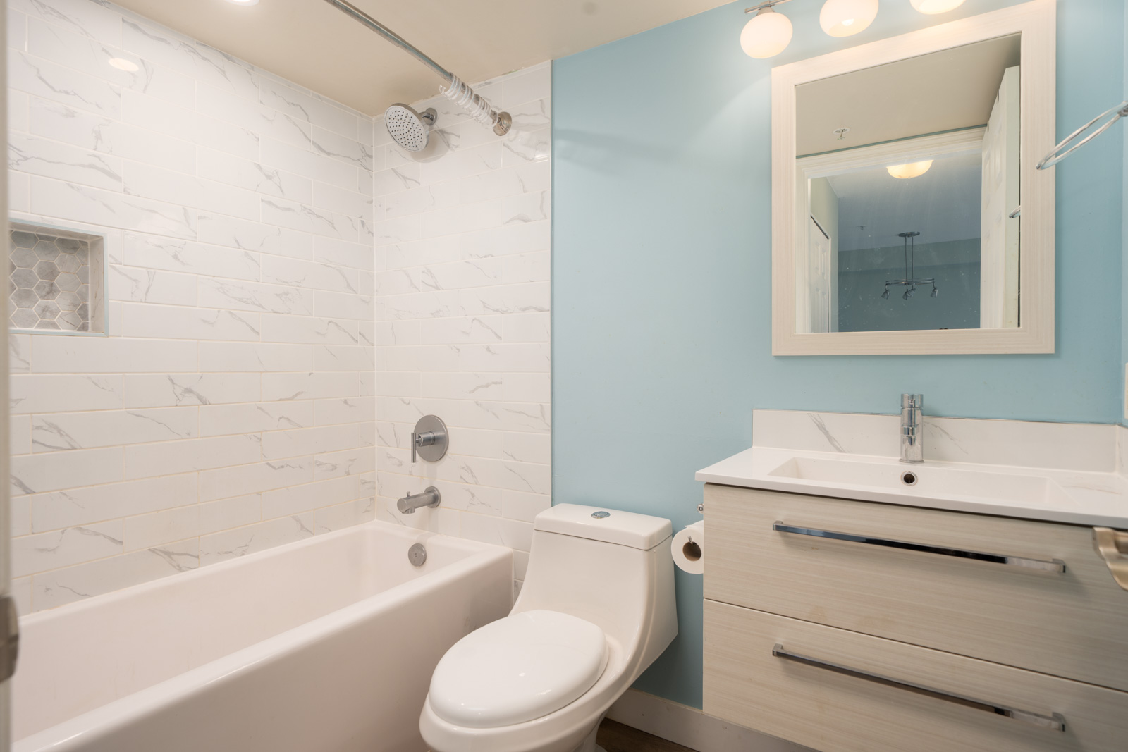 Modern bathroom at 1099 E Broadway, Vancouver with a white tub, vanity, blue accent wall, tiled shower with shelf, and three mirror lights.