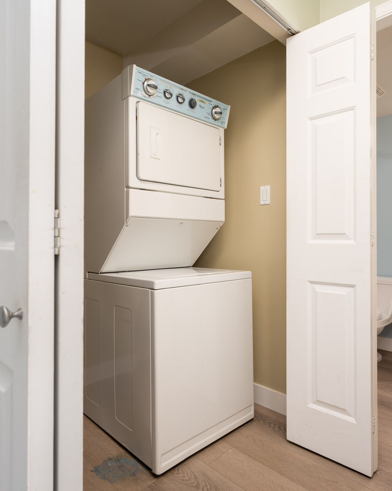 Stacked washer and dryer in a small closet with folding doors, beige walls, and wood floors on the 3rd floor at 1099 E Broadway, Vancouver.