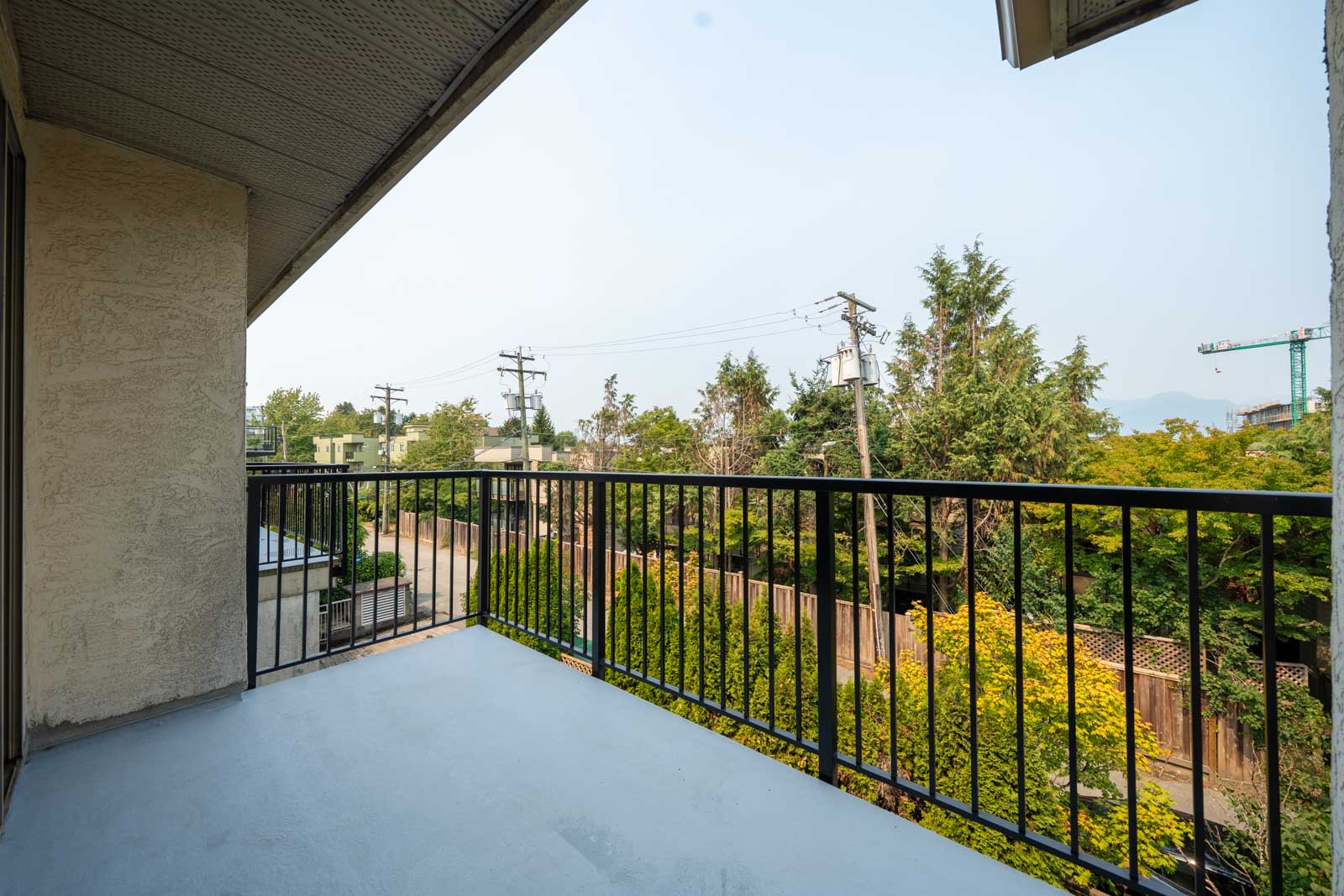 View from a concrete balcony with black metal railing, looking over trees, power lines, and a quiet street in Vancouver near 1099 E Broadway.