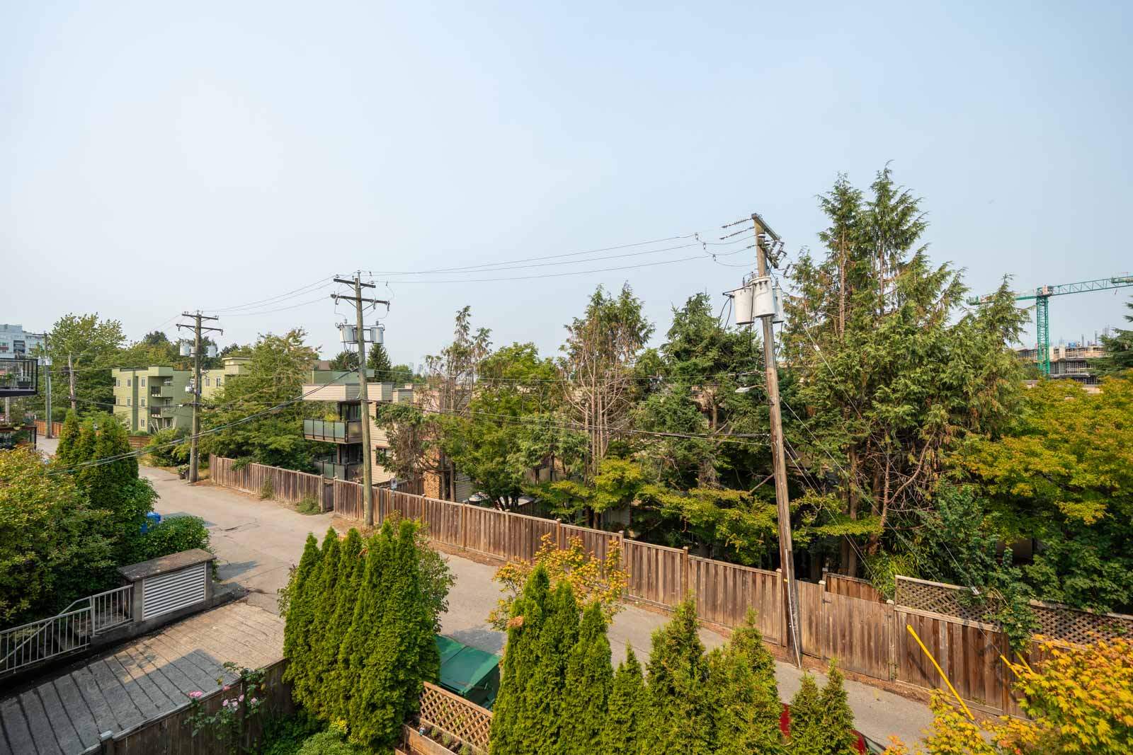Vancouver residential street with wooden fence, trees, utility poles, and power lines; buildings and crane in background near 1099 E Broadway.