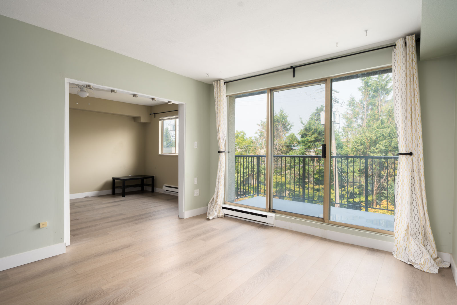 Bright, empty living room at 1099 E Broadway, Vancouver with light wood floors, pale green walls, large windows, and tree views.