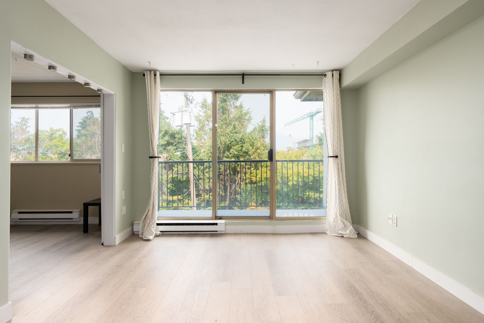 Empty room with light wood floors, pale green walls, large sliding doors, white curtains, and a balcony view in Vancouver.