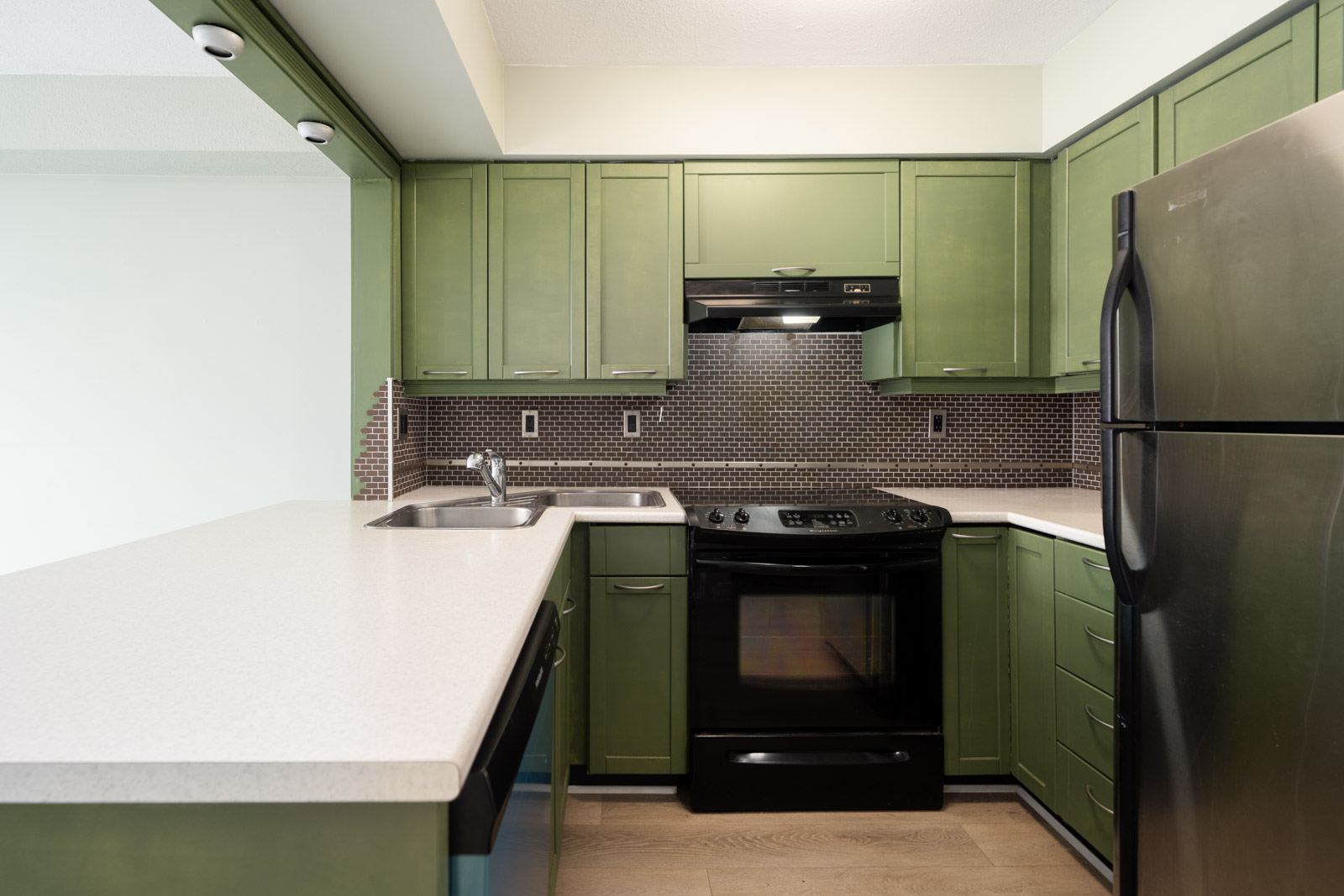 Modern kitchen at 1099 E Broadway, Vancouver, with green cabinets, black appliances, white counters, and brown tile backsplash.