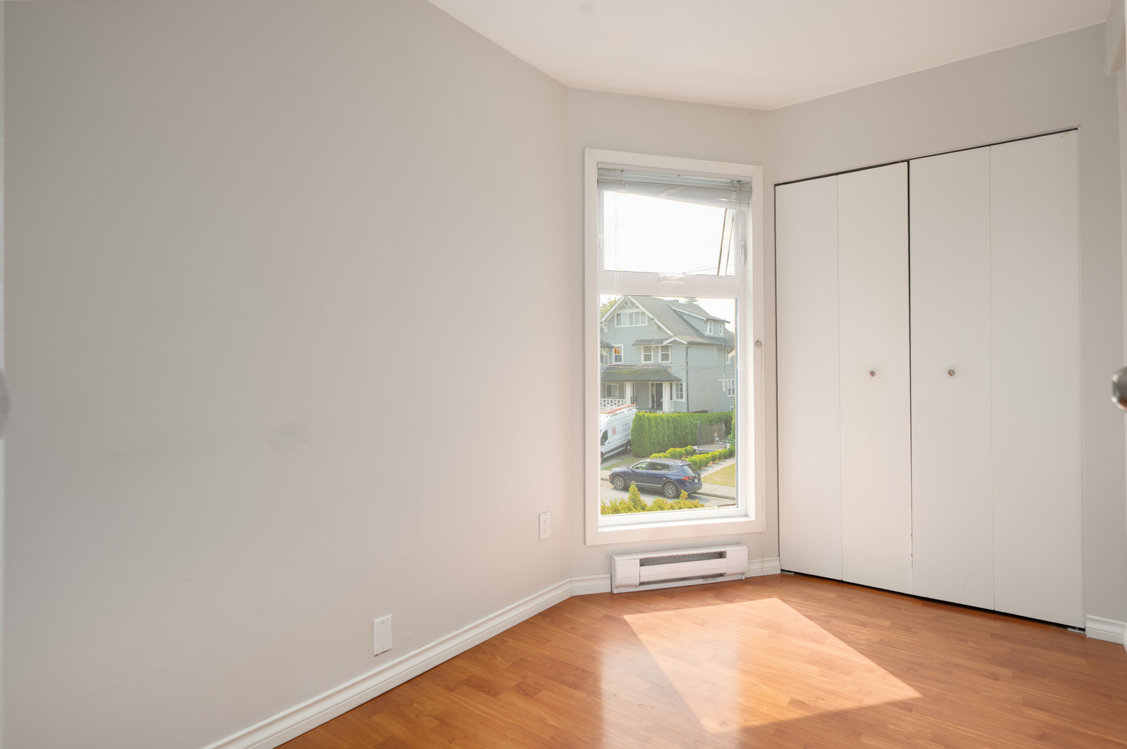 Empty room with light gray walls, large window facing a house and car, closed white closet, and light wood floor in Vancouver.