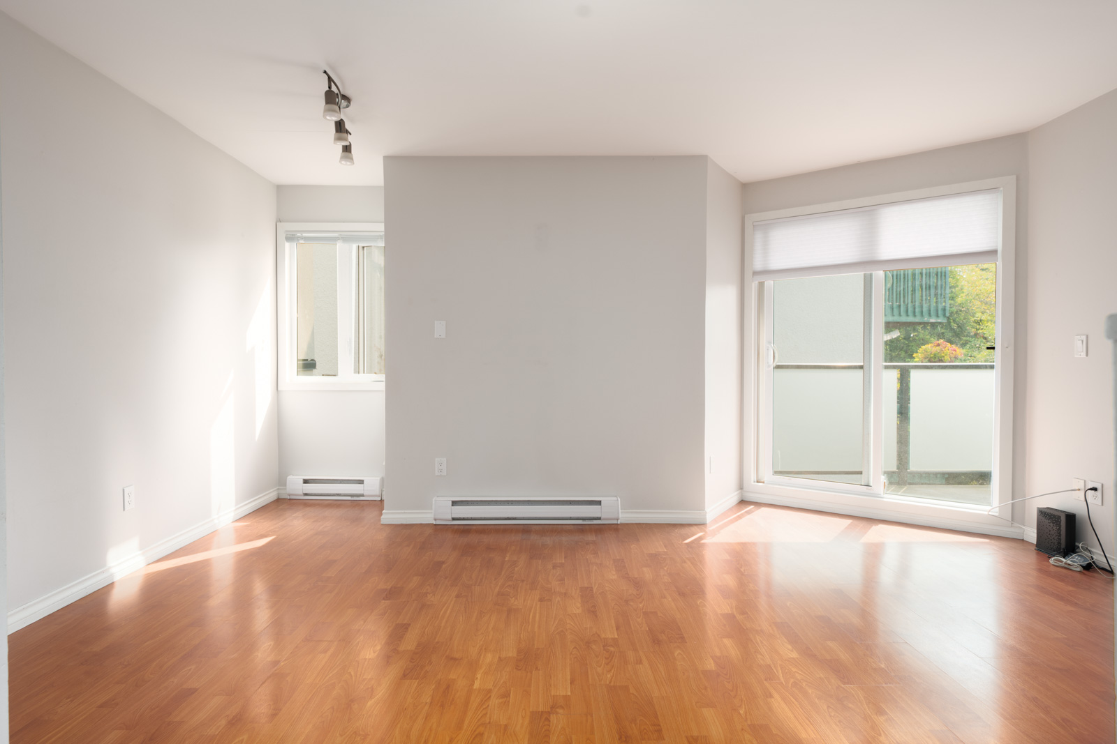 Empty 2nd floor room with light walls, wood laminate floors, track lighting, two large windows, and baseboard heater in Vancouver.