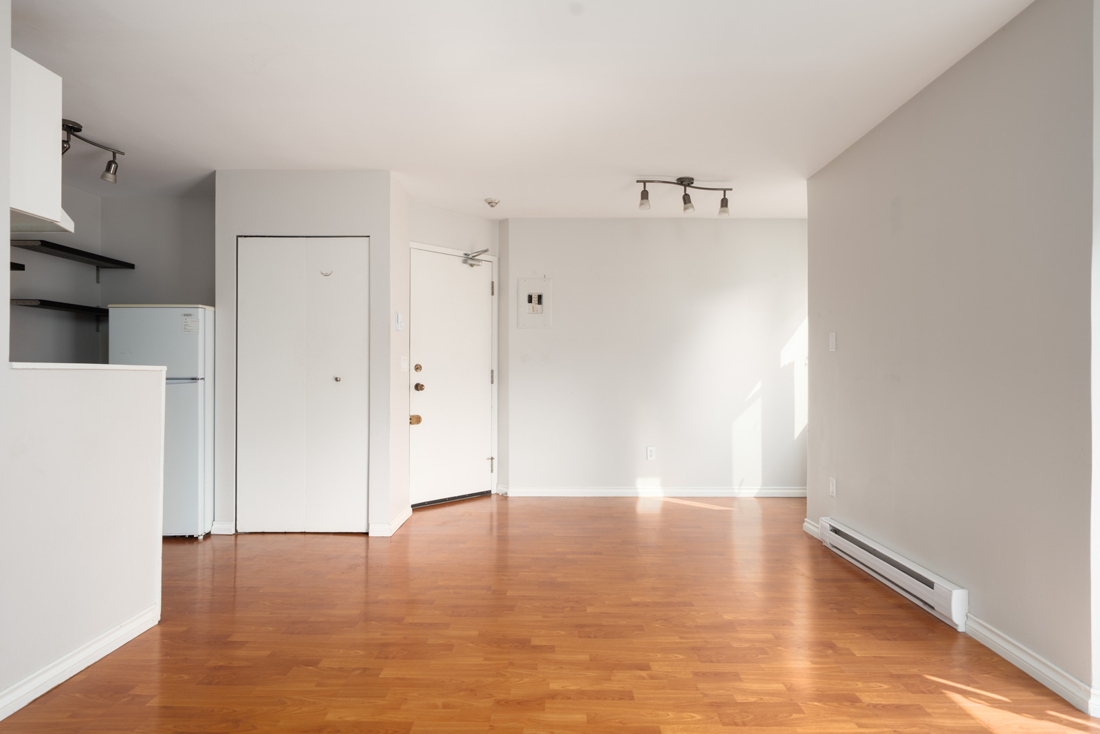 Empty room with light wood floors, white walls, closed door, small corner fridge, and ceiling lights on 2nd floor in Vancouver.