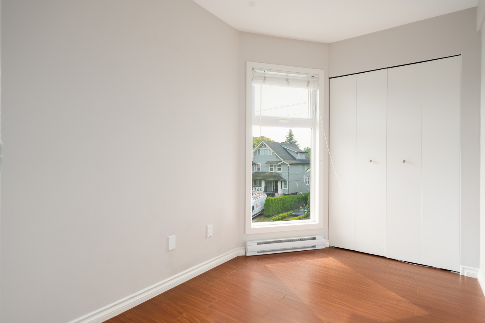 Empty room with light gray walls, wood flooring, white closet doors, and a large window overlooking trees in Vancouver.