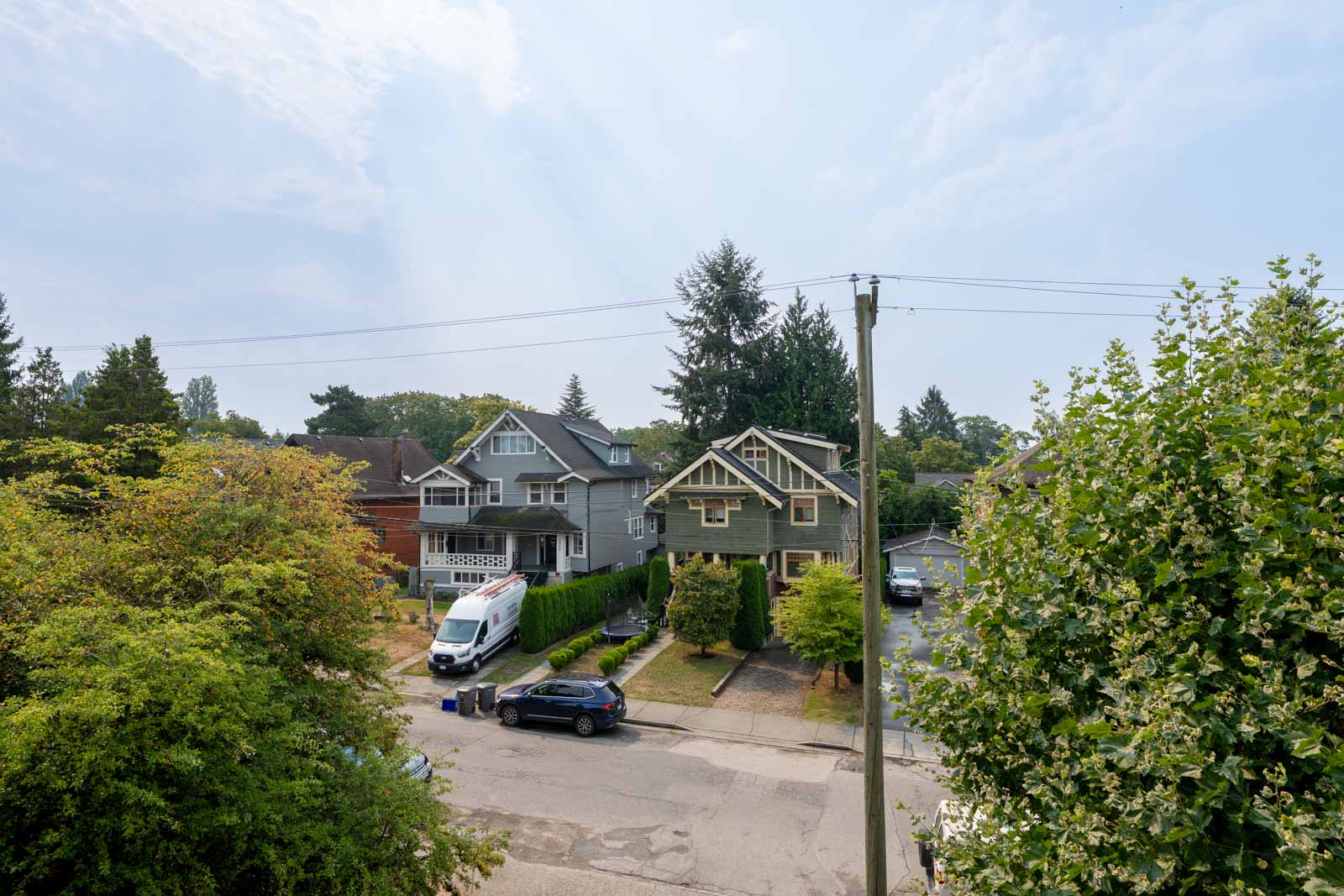 Residential street in Vancouver with houses, trees, a white van in a driveway, and a black car near a utility pole under clouds.