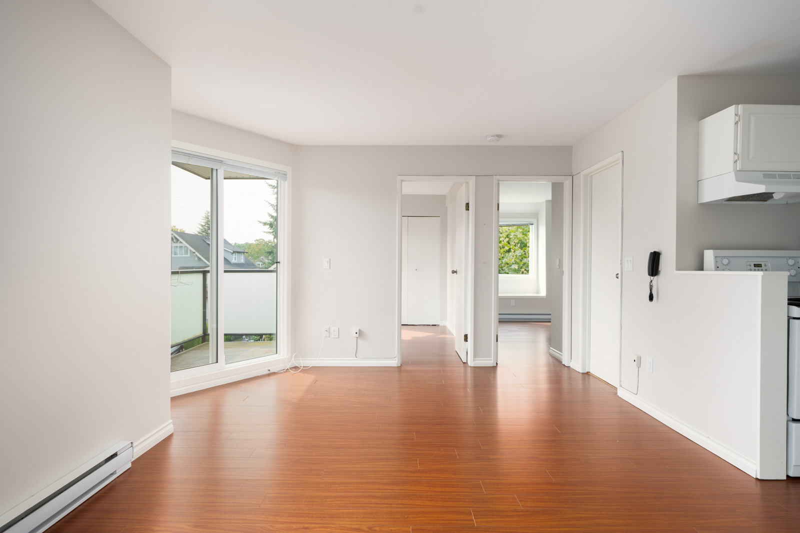 Unfurnished apartment room in Vancouver with light walls, wood floors, and a sliding glass door to a 3rd-floor balcony; kitchen on right.
