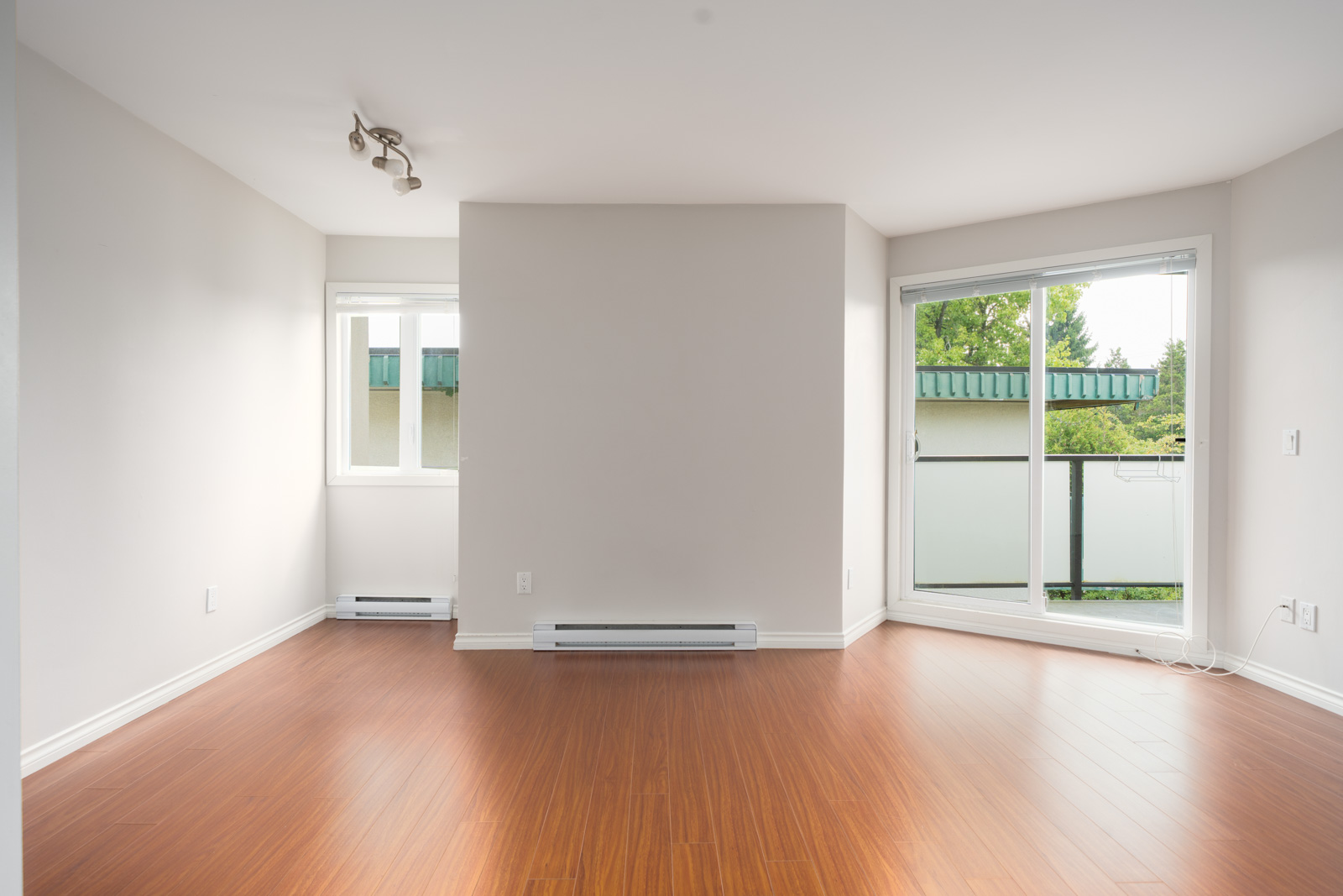 Empty room on 3rd floor, West 5th Ave, Vancouver, with light walls, wood floors, window, sliding door to balcony with greenery.