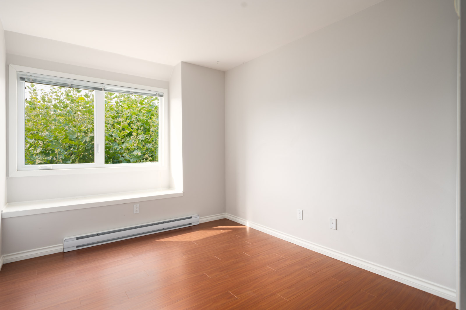 Empty room on 3rd floor at 2365 West 5th Ave, Vancouver, with light walls, big window overlooking trees, heater, and wood floors.