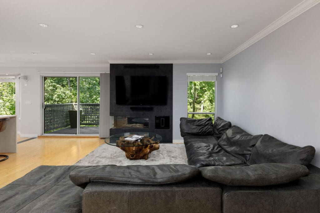 Modern Vancouver living room with black sectional, glass coffee table, wall TV, fireplace, light wood floors, and tree views.