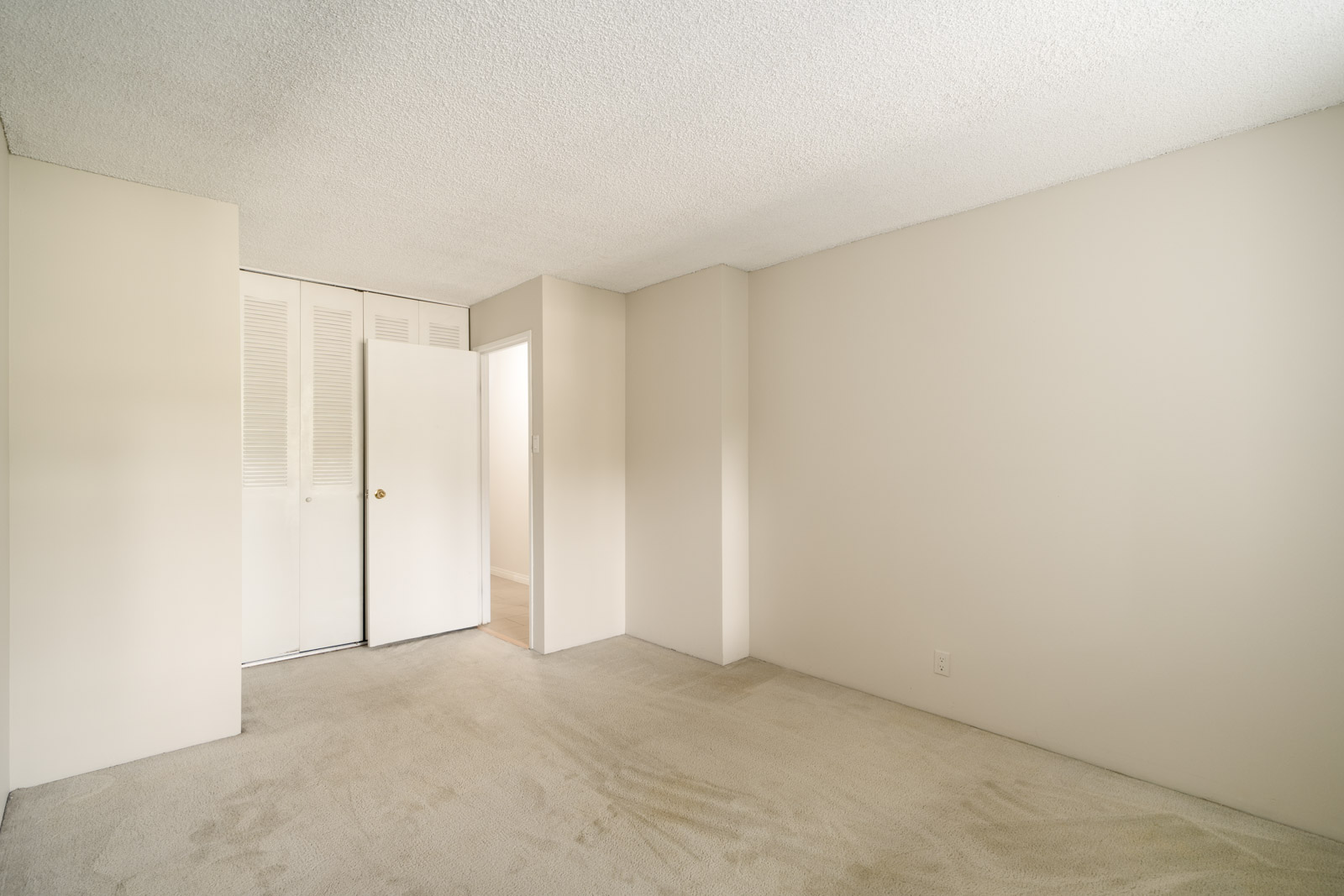 Empty beige carpeted room on Fullerton Ave, Vancouver, with shut closet doors, one open door, and plain off-white walls and ceiling.
