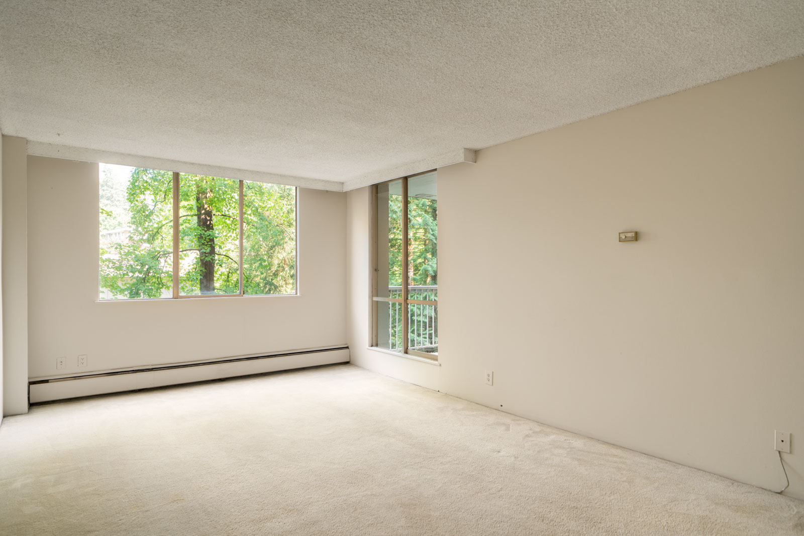 Empty room with beige carpet, off-white walls, large window, sliding door to balcony, baseboard heater; trees outside on Fullerton Ave, Vancouver.