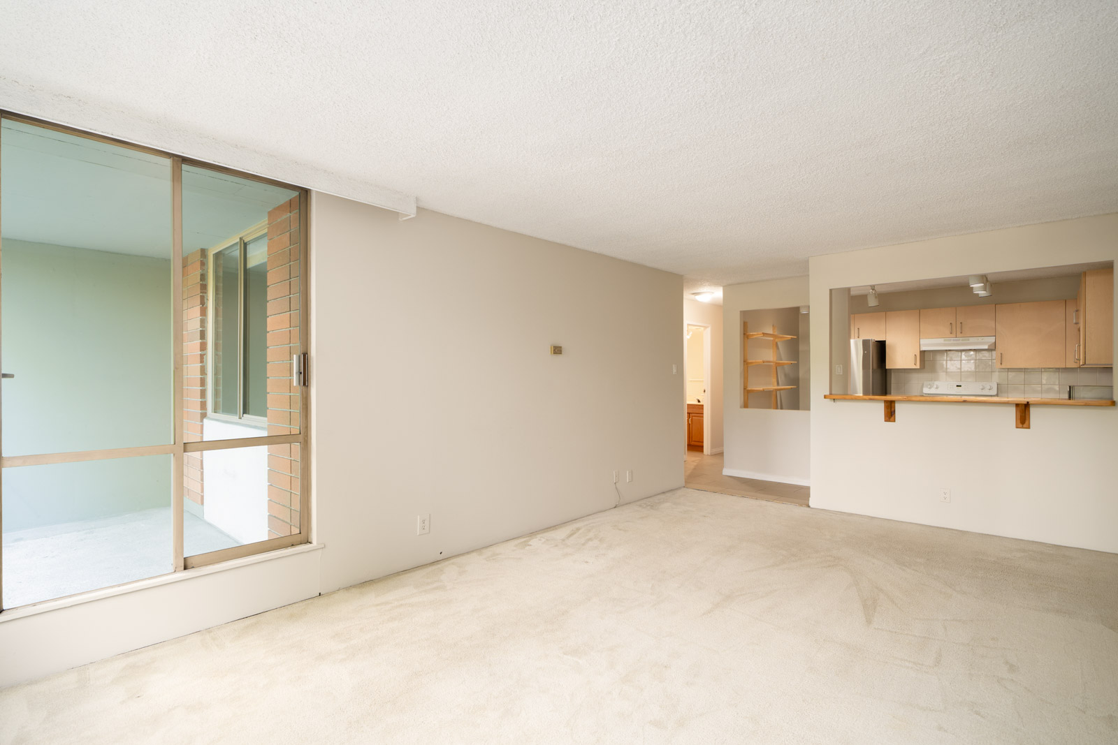 Unfurnished Vancouver apartment with carpeted floors, large window to enclosed balcony, and open kitchen with light wood cabinets.