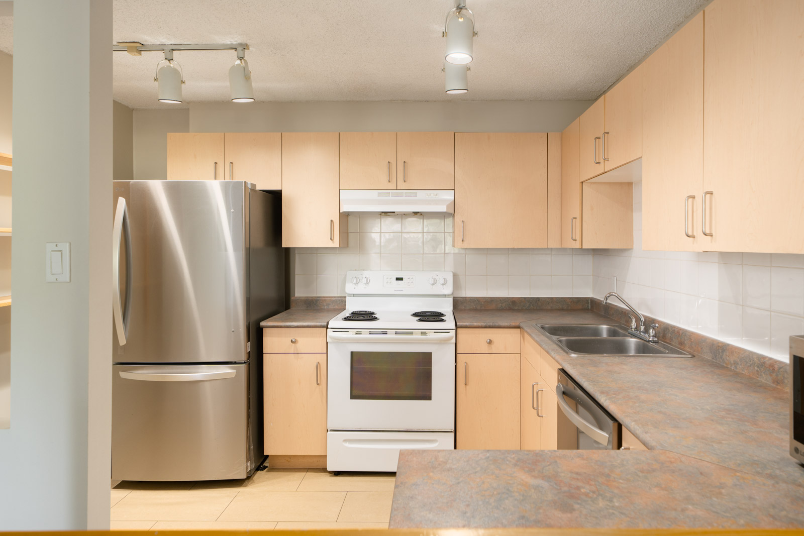 Modern Vancouver kitchen with light wood cabinets, stainless fridge, white stove, double sink, and neutral-toned counters.