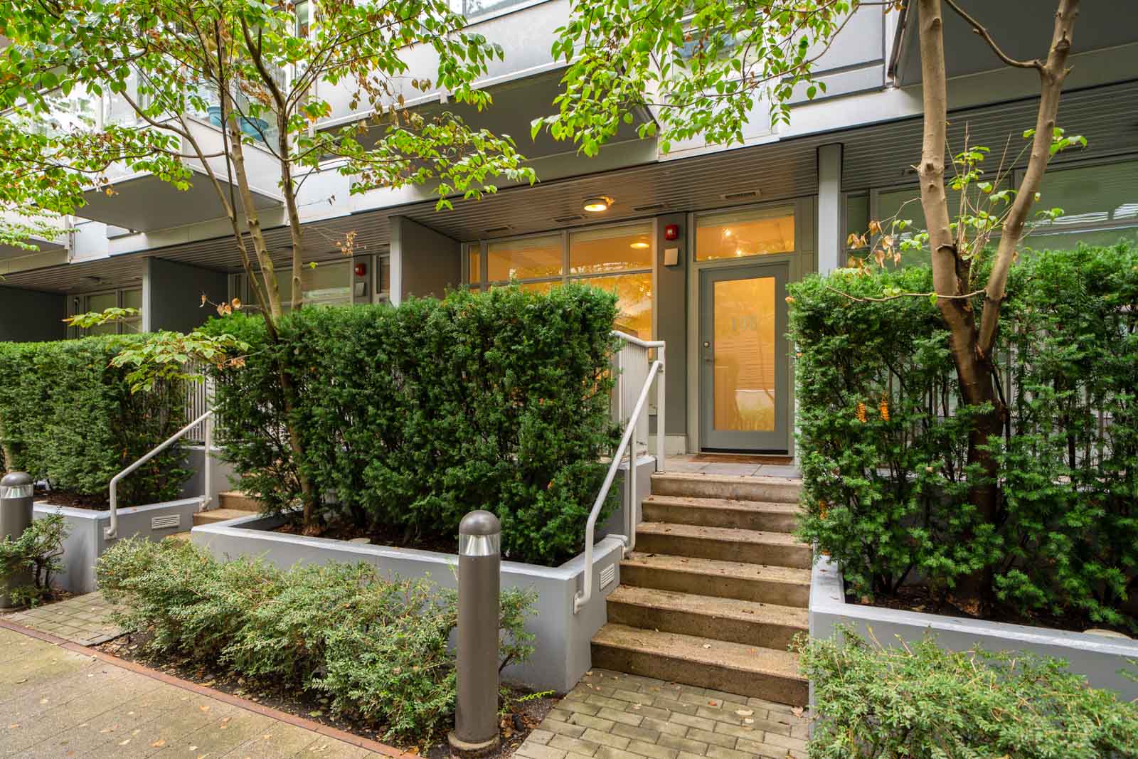 Modern townhouse entrance in Vancouver with stairs, glass door, large windows, and trimmed hedges along the sidewalk.