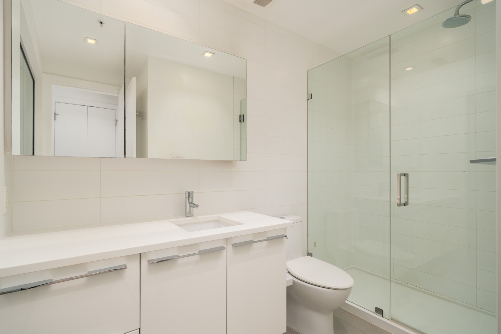 Modern bathroom at 108 E 1st Ave, Vancouver with white cabinets, countertop sink, mirror, toilet, and glass shower with tiled walls.