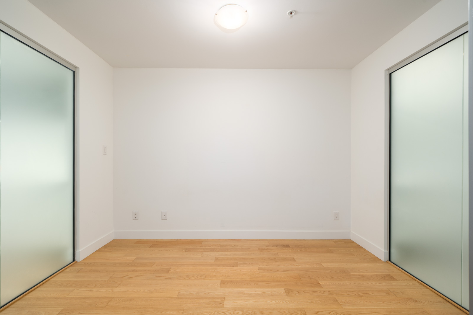 Empty room with light wood floors, white walls, ceiling light, and two frosted glass sliding doors in a Vancouver townhouse.