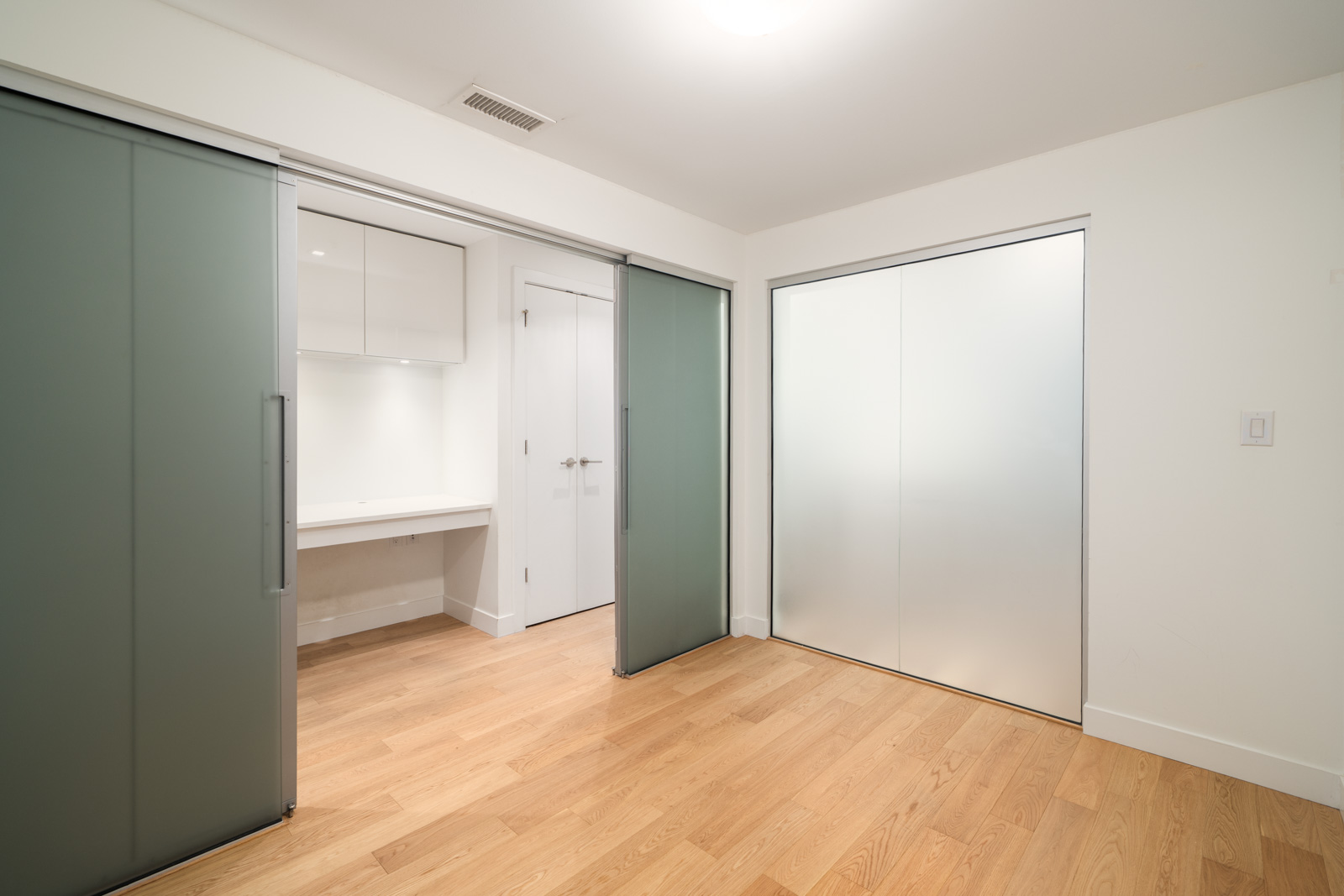 Empty room with light wood flooring, white walls, sliding frosted closet doors, and built-in desk with cabinets in Vancouver townhouse.
