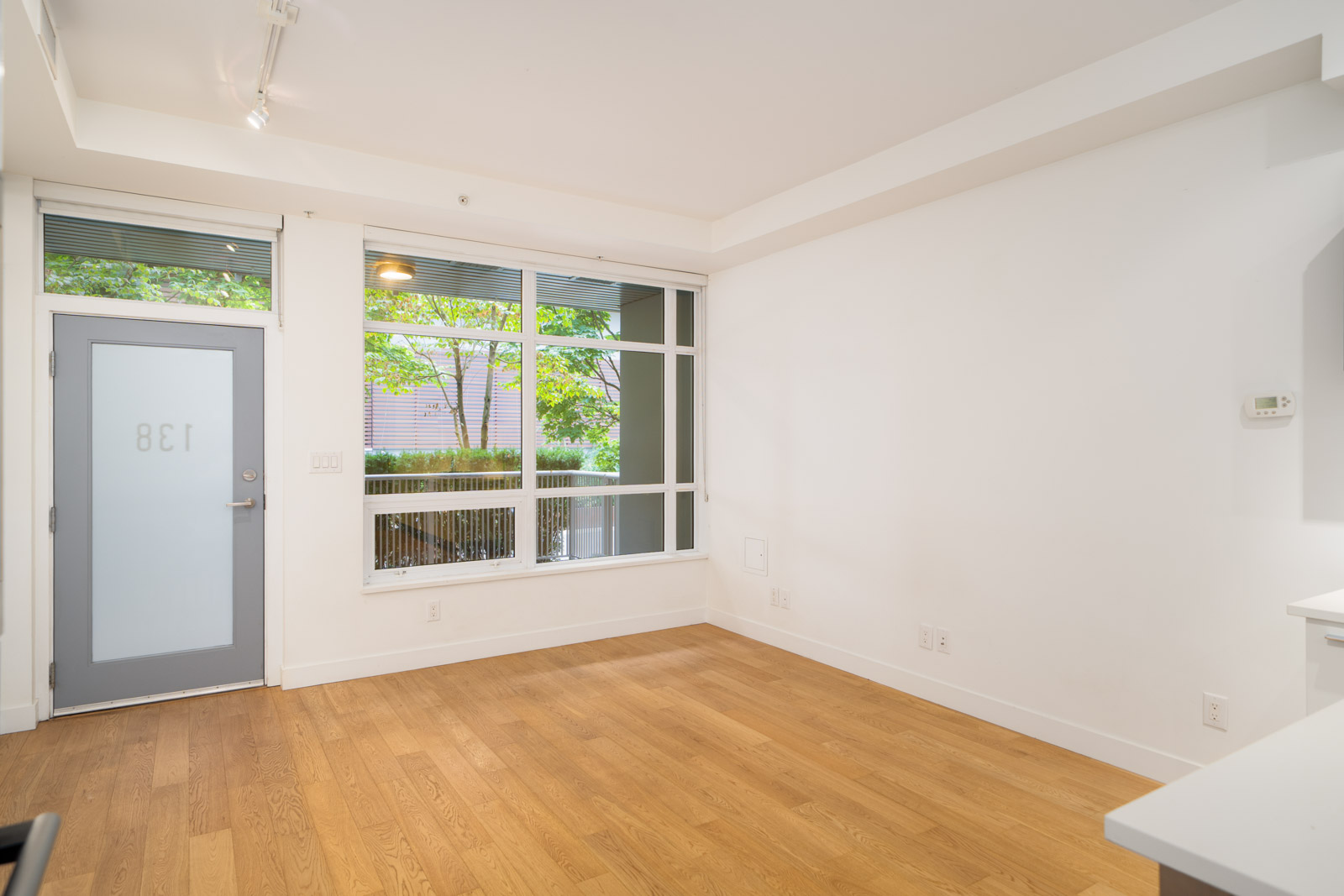 Unfurnished room with light wood floors, white walls, large window, gray door marked 138; trees and fence outside in Vancouver.