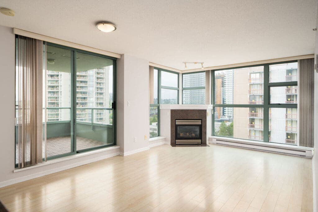 Bright, empty living room with light wood floors, large windows, balcony access, and fireplace in a 16th floor Burnaby condo.