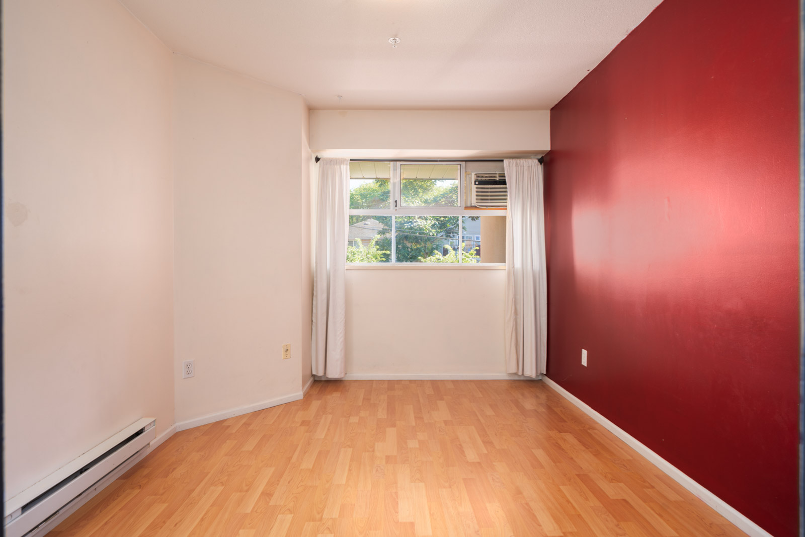 Empty room with light wood floors, a red accent wall, cream walls, white-curtained window, and baseboard heater in Vancouver.