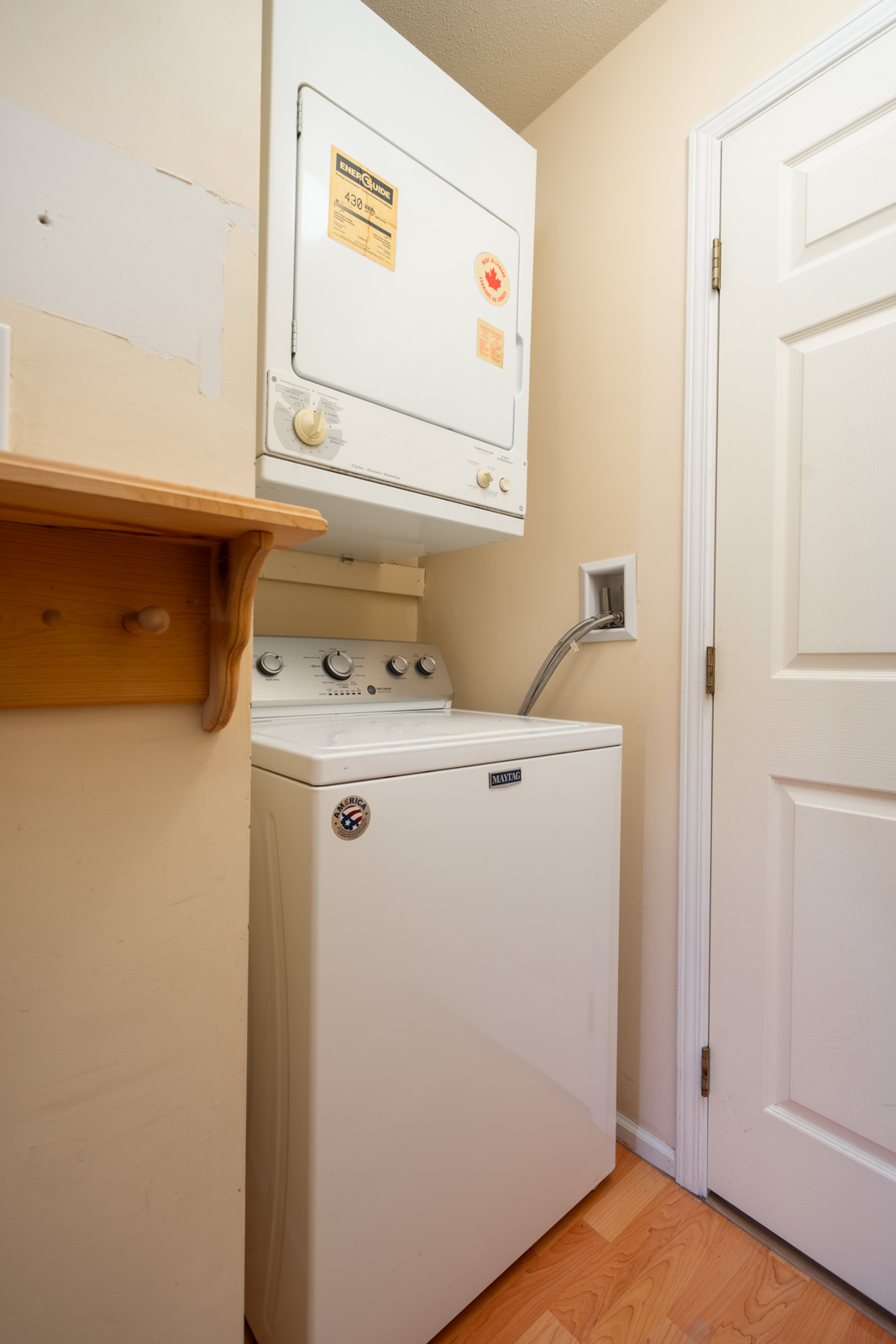 A stacked washer and dryer in a compact laundry room with light walls, wood flooring, white door, and wooden shelf in Vancouver.