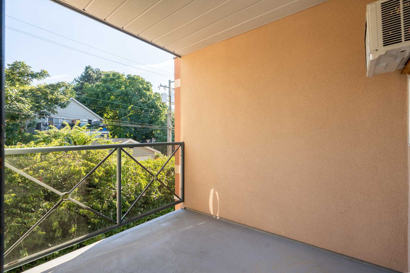 Empty 3rd-floor balcony with beige wall, metal railing, AC unit, overlooking trees and power lines on Triumph St in Vancouver.