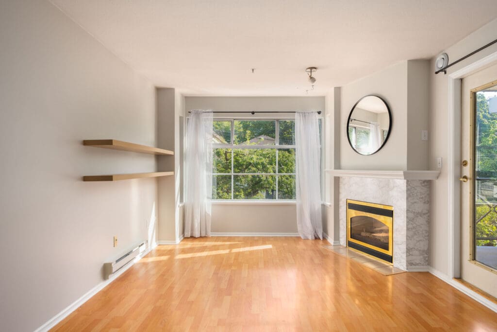 Bright empty room with light wood floors, large window with sheer curtains, wall shelves, and a corner fireplace with round mirror in Vancouver.