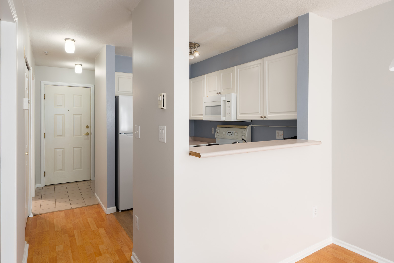 Small kitchen with white cabinets, light wood floors, and a hallway to a white front door in a Triumph St, Vancouver home.