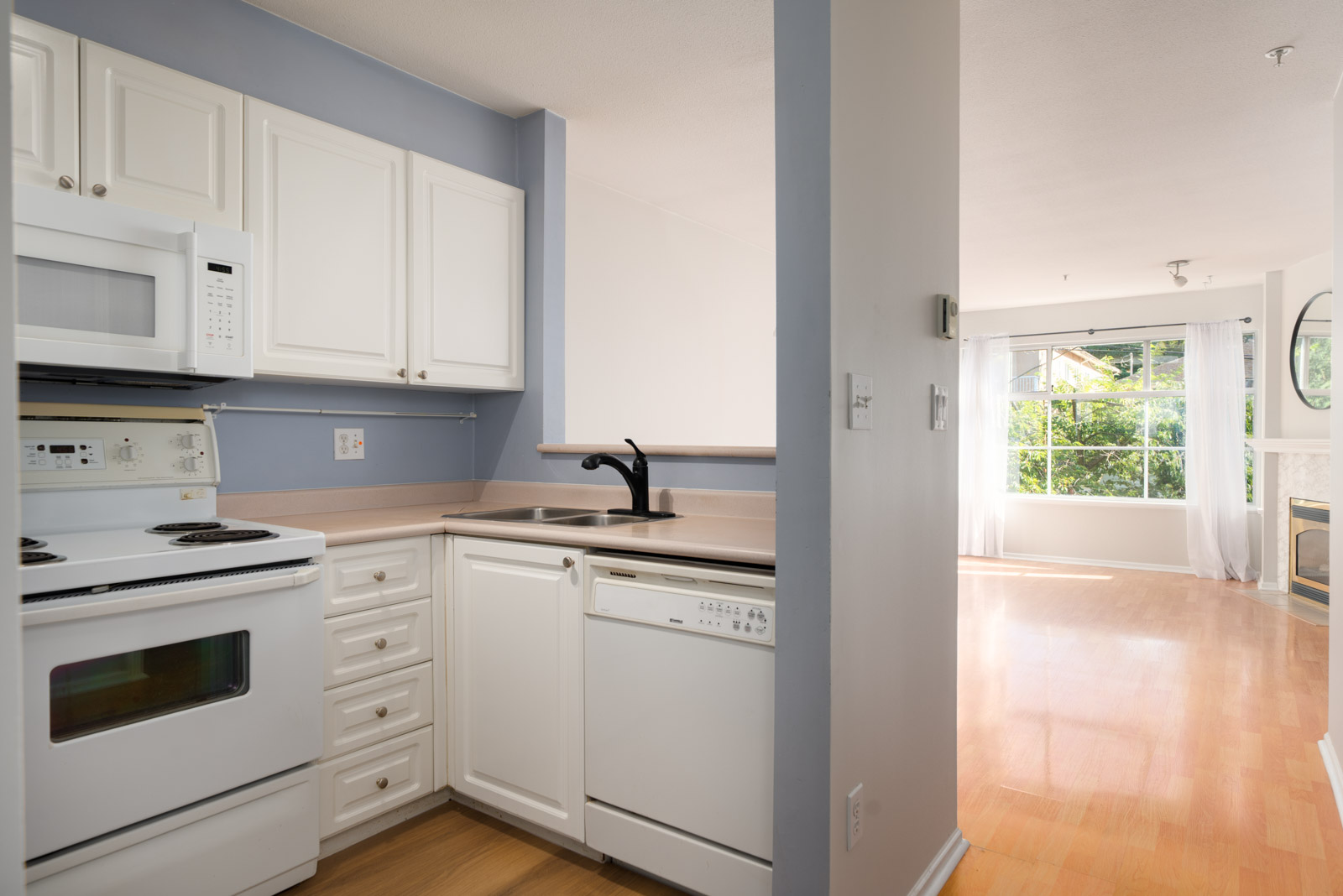 White-cabinet kitchen with appliances and sink beside living area with large windows, wood floor; Triumph St, Vancouver.