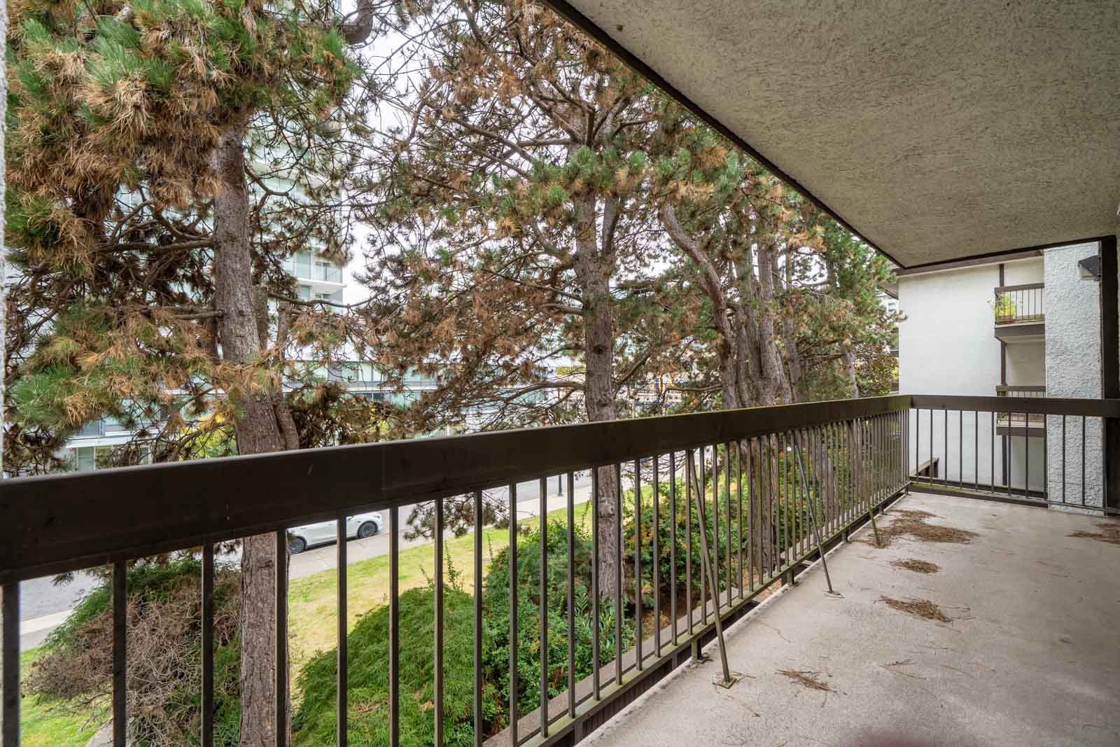 Covered 2nd floor balcony with metal railing overlooks trees, bushes, parked cars on Buswell Street, Richmond, on a cloudy day.