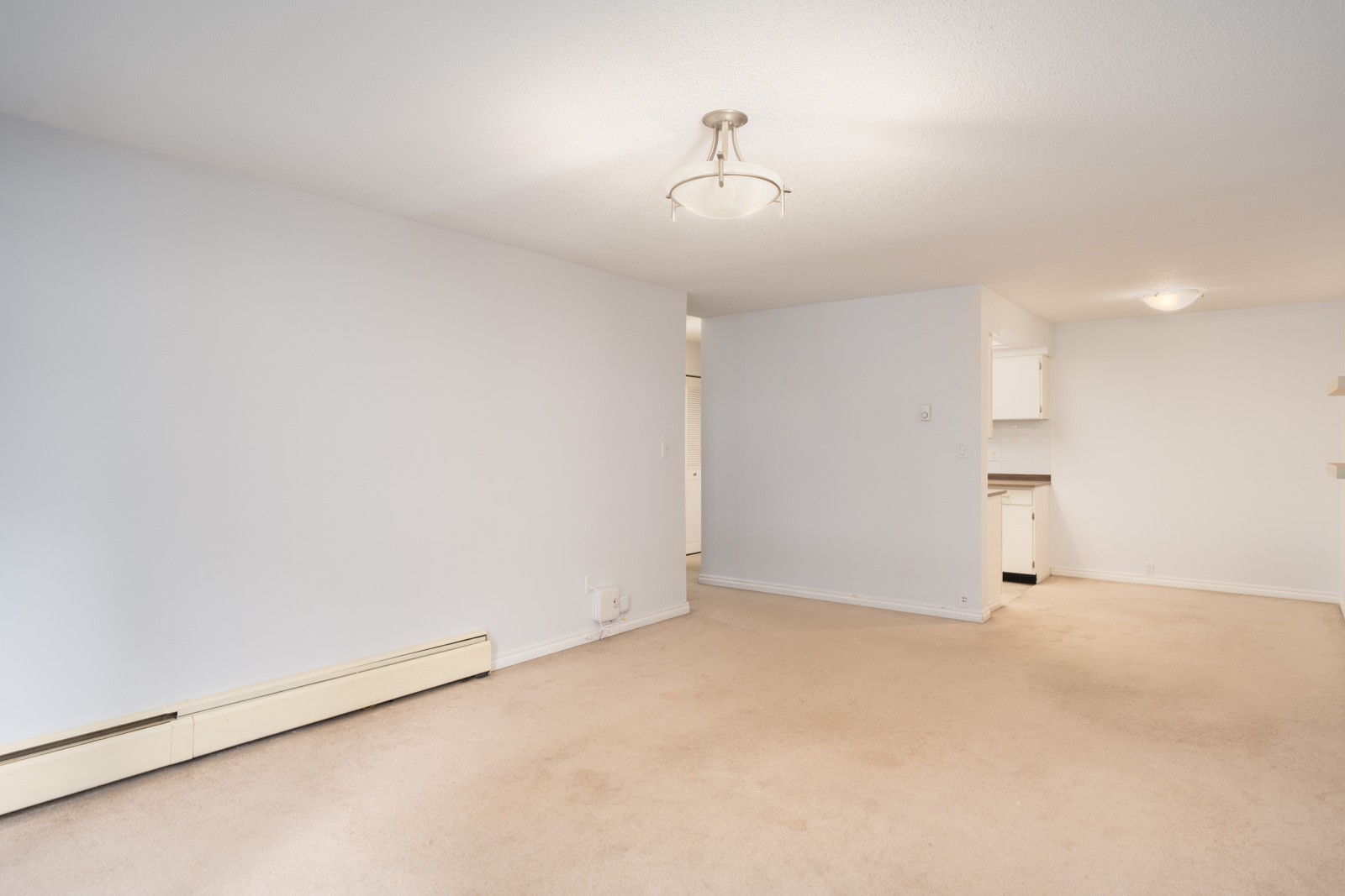 Unfurnished room at 6560 Buswell, Richmond with beige carpet, white walls, ceiling light, heater, and small kitchen in background.