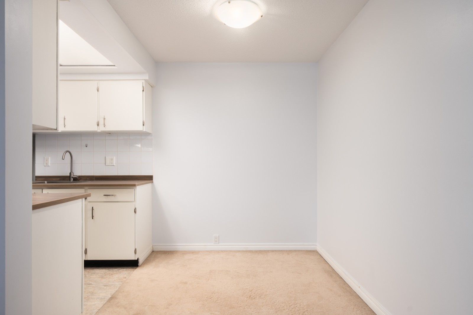 Empty room with beige carpet, white walls, and a kitchen area with white cabinets on Buswell Street in Richmond; ceiling light above.