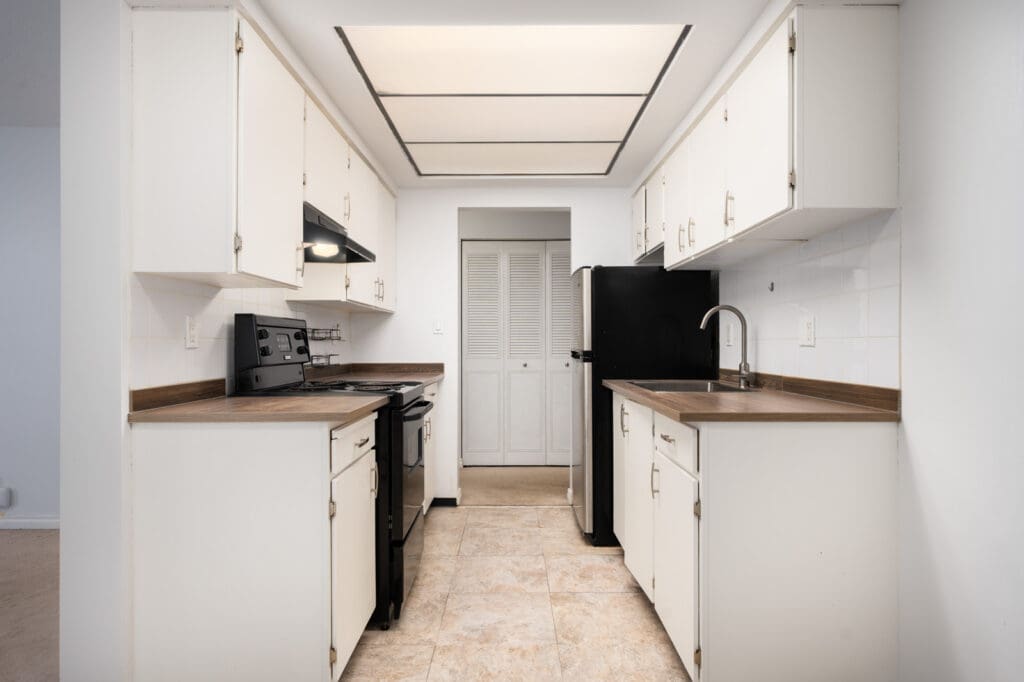 Galley kitchen with white cabinets, brown counters, black appliances, tile floor, ceiling light, and door with white shutters. Richmond, 2nd floor.