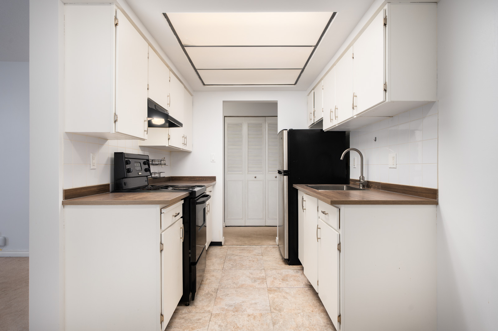 Galley kitchen with white cabinets, brown counters, black appliances, tile floor, ceiling light, and door with white shutters. Richmond, 2nd floor.