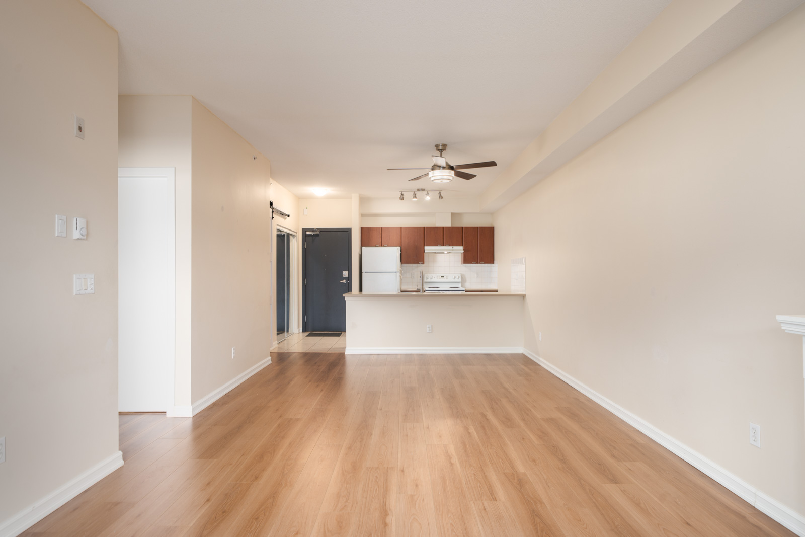 Empty apartment with light wood floors, beige walls, open layout to kitchen with white appliances and ceiling fan near 148 Street, Surrey.